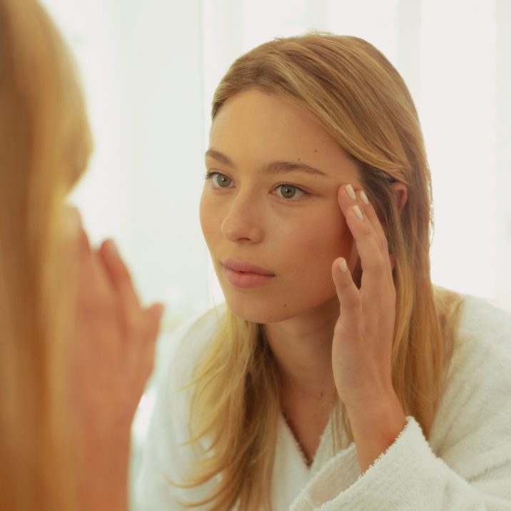Woman in a white robe looking in a mirror and touching her temple