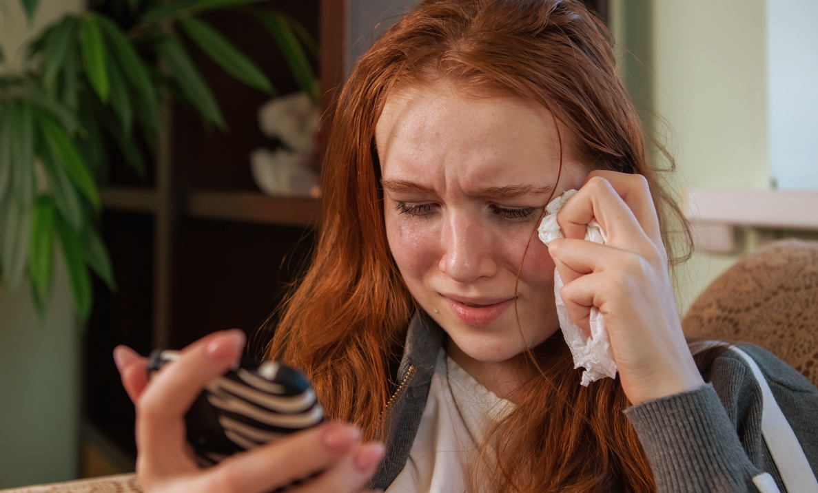 Young woman with red hair wiping her tearful eye with a tissue while looking at a hand mirror
